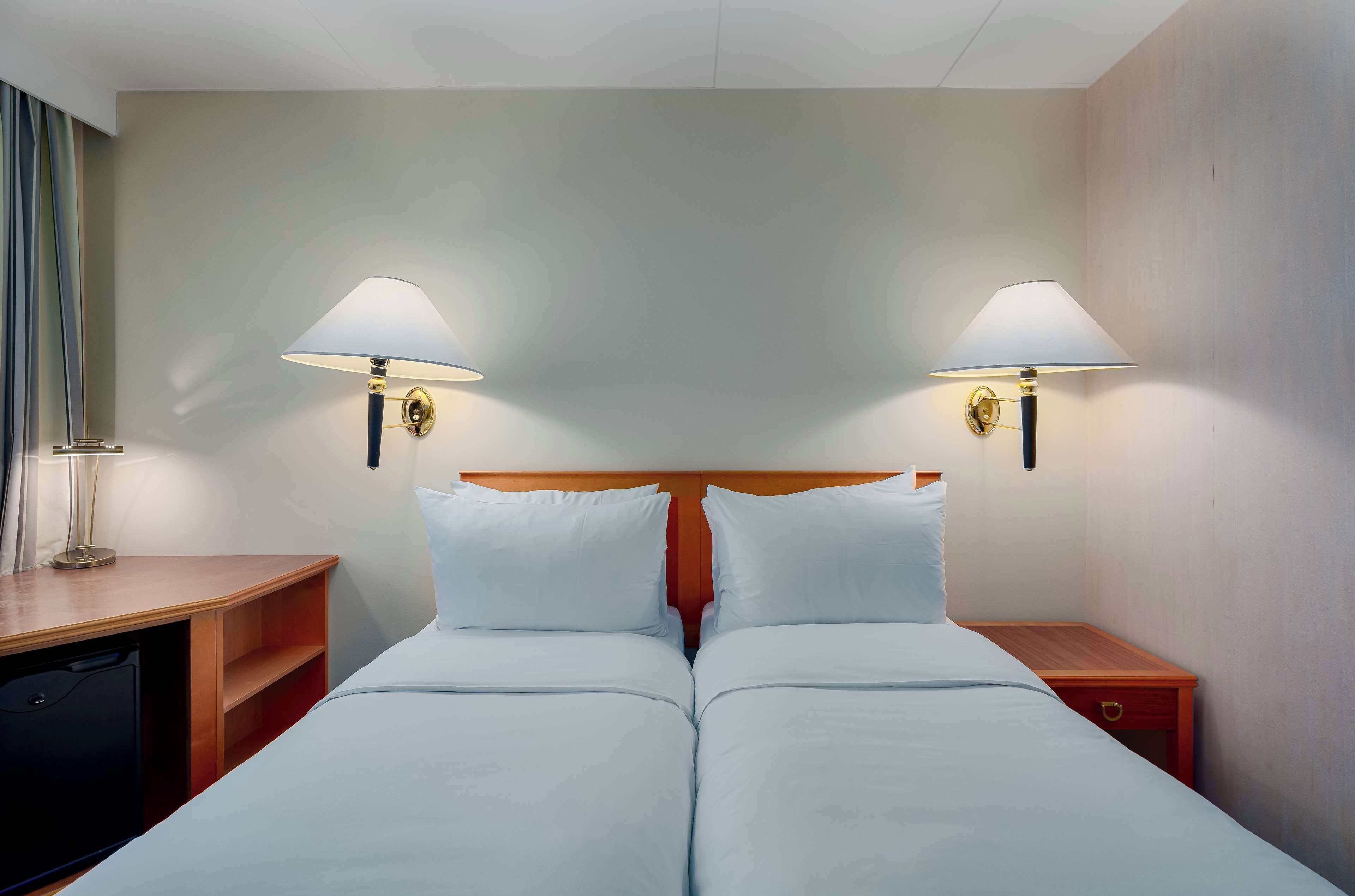 Quarto de hotel com duas camas, cabeceira de madeira, mesas de cabeceira e luminárias fixadas na parede.