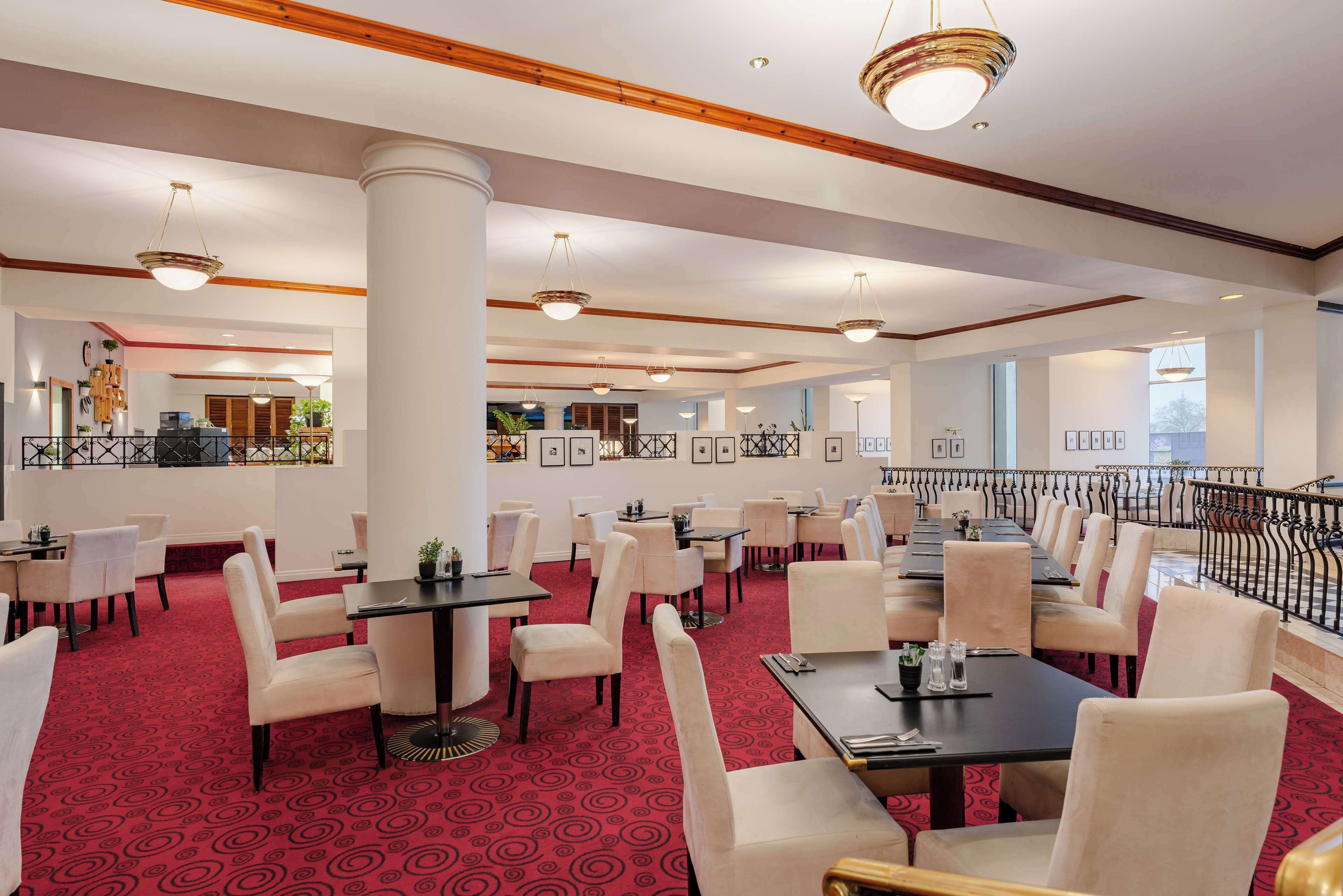 Un hall de hotel con varias mesas y sillas para comer, piso cubierto de alfombra roja y un balcón con barandilla.
