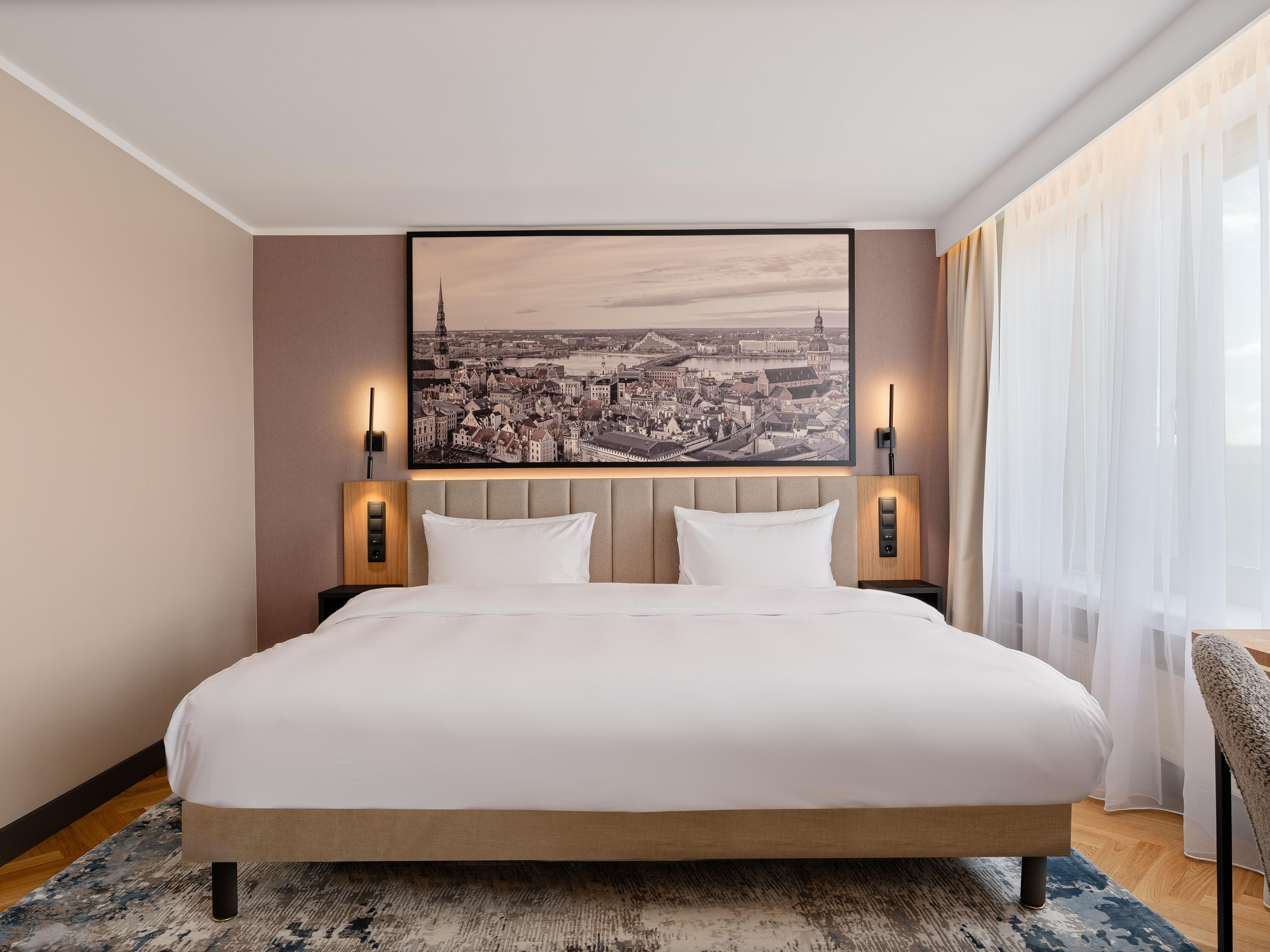 A hotel room with a large bed, two bedside lamps, a cityscape painting on the wall, and a window with sheer curtains.