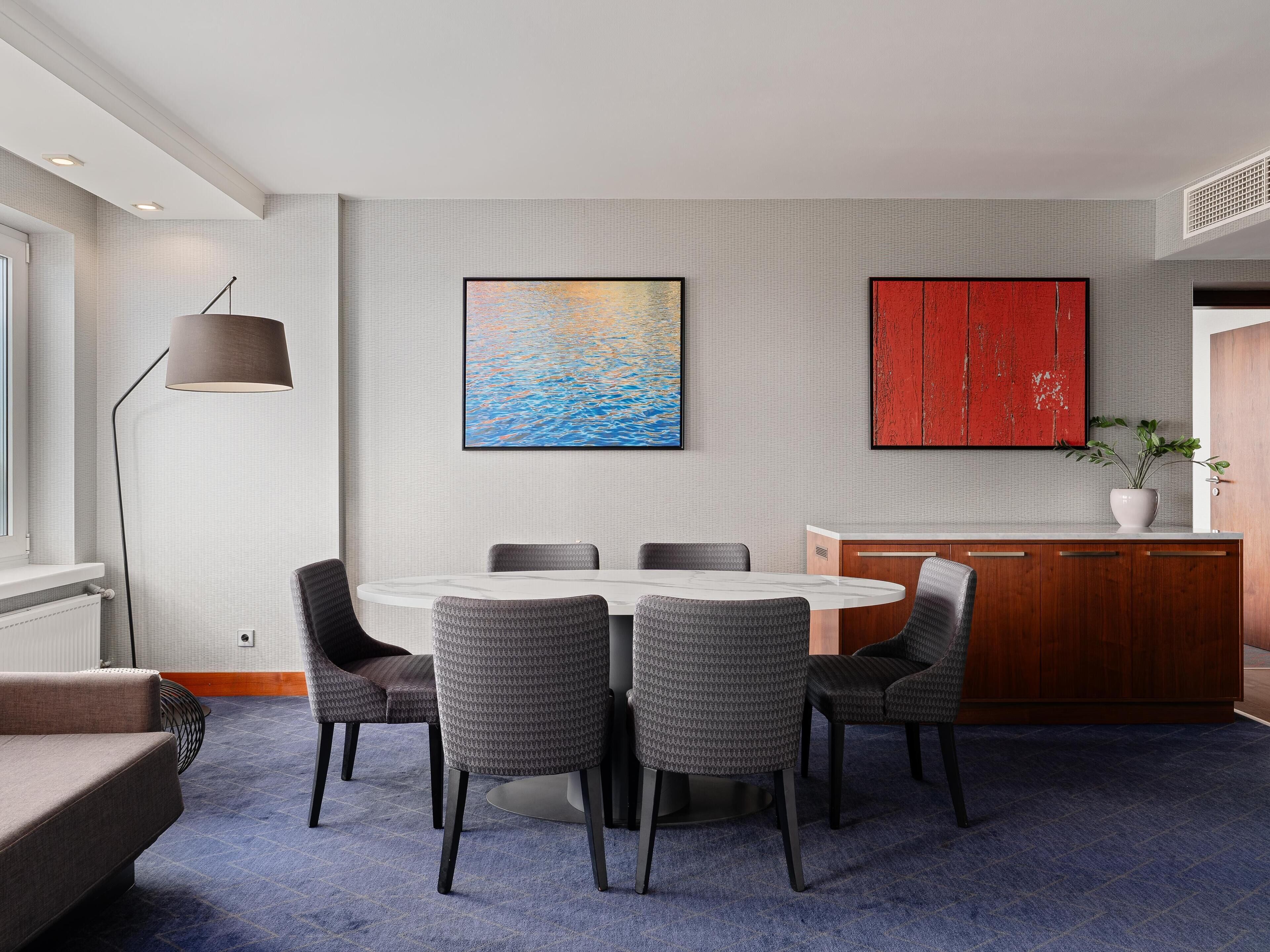 A modern dining area with a round table, grey upholstered chairs, a pendant lamp, and a red cabinet.
