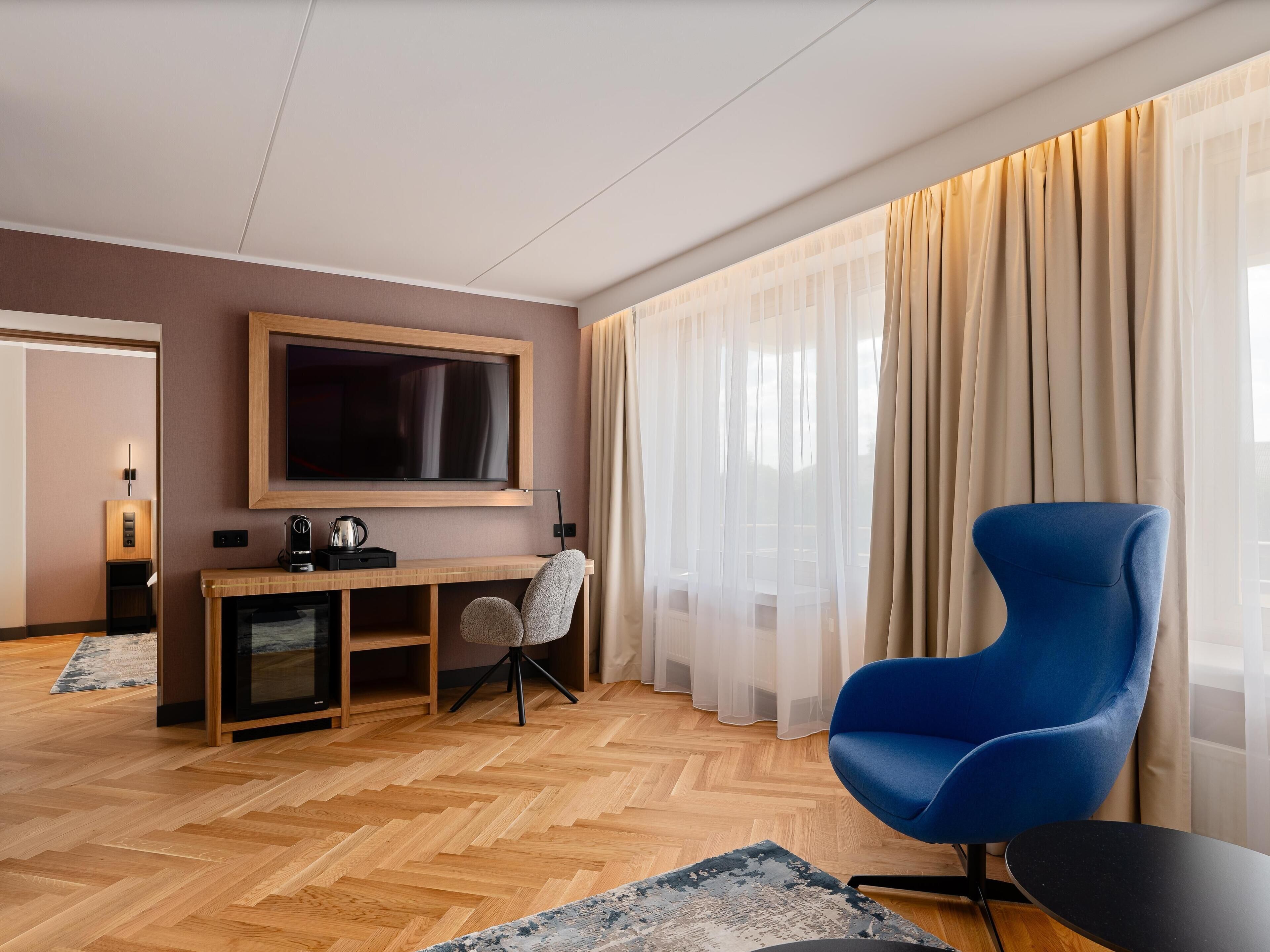 Moderni hotellihuone, jossa on puinen kalanruotokuvioinen lattia, suuri seinälle asennettu taulutelevisio, sininen nojatuoli, pieni työpöytä kahvinkeittimellä sekä läpikuultavat verhot.