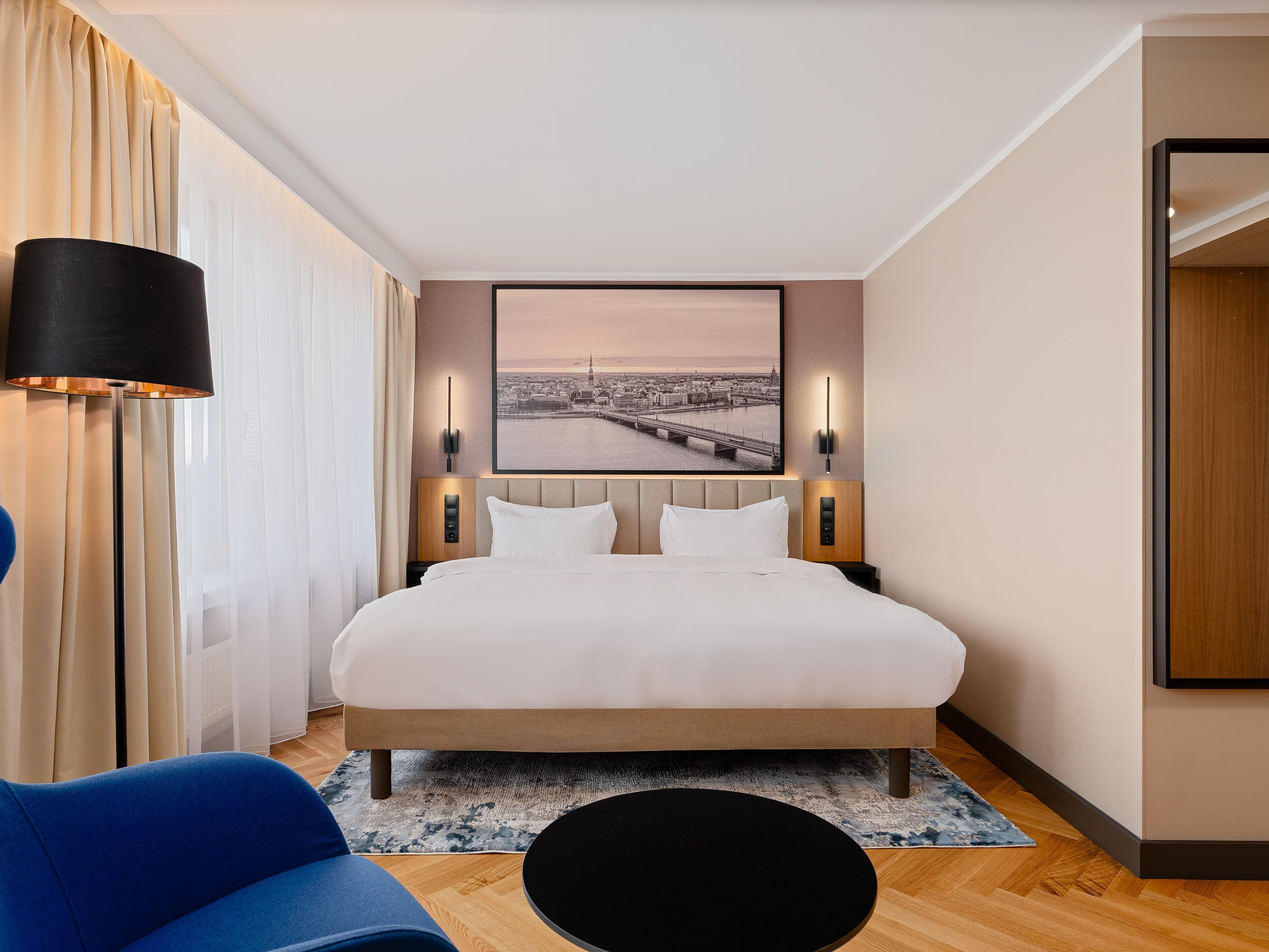 Una habitación de hotel con una cama, una mesita de noche, una lámpara, una silla, una alfombra y un cuadro colgado en la pared.