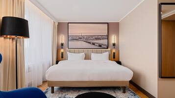 Una habitación de hotel con una cama, una mesita de noche, una lámpara, una silla, una alfombra y un cuadro colgado en la pared.