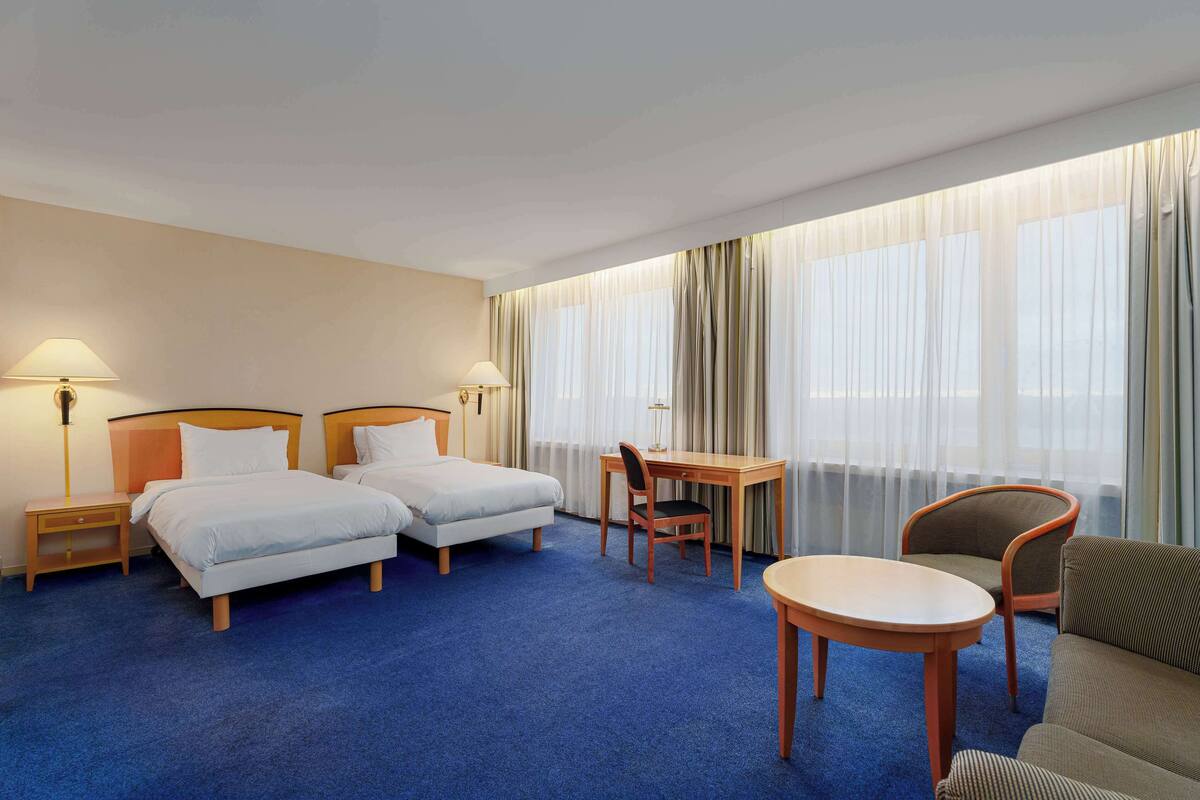A hotel room with two beds, a desk, a chair, a sofa, and large windows with sheer curtains.