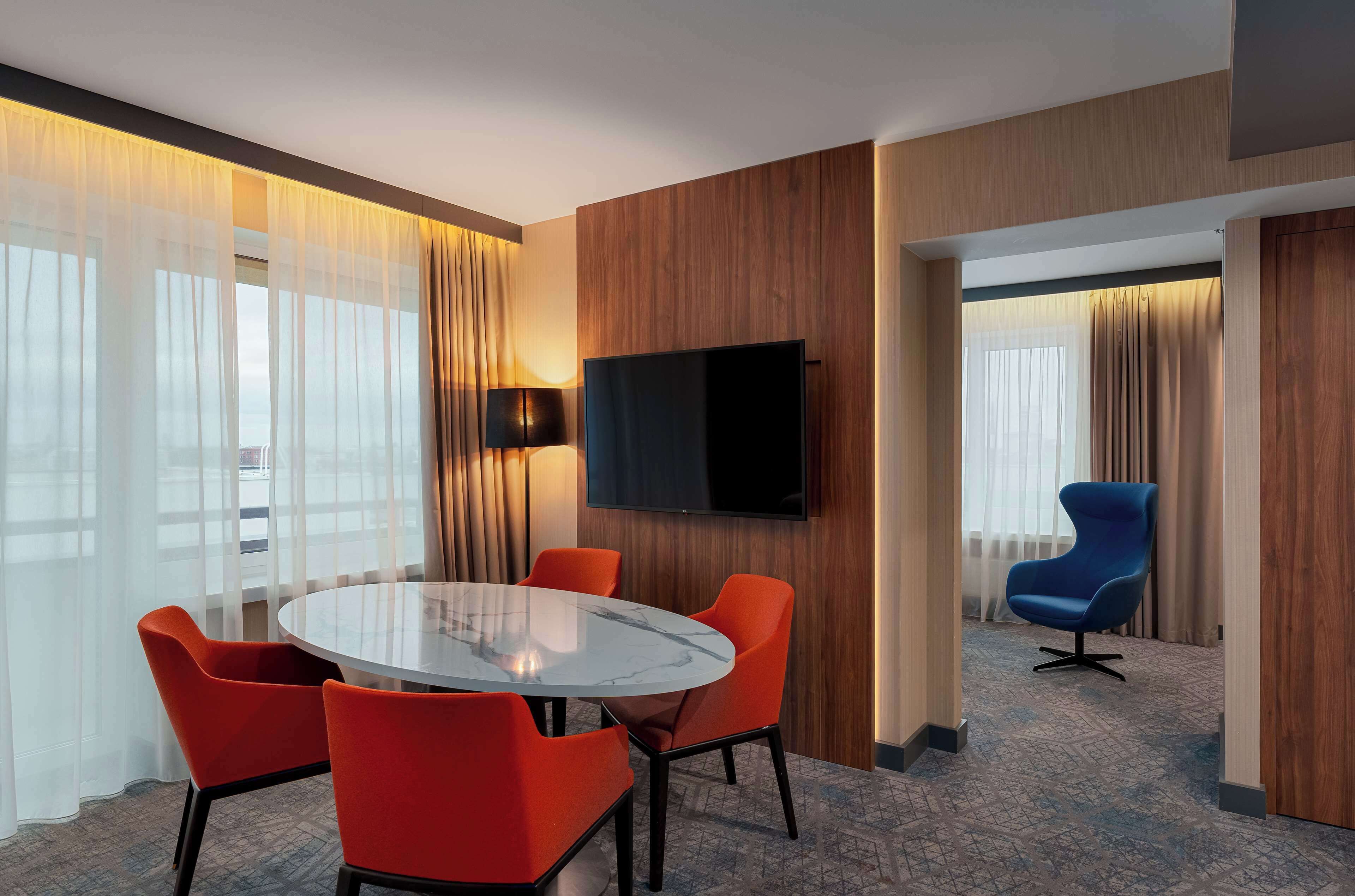 Habitación de hotel con una mesa redonda, sillas anaranjadas, un televisor de pantalla plana y un sillón azul.