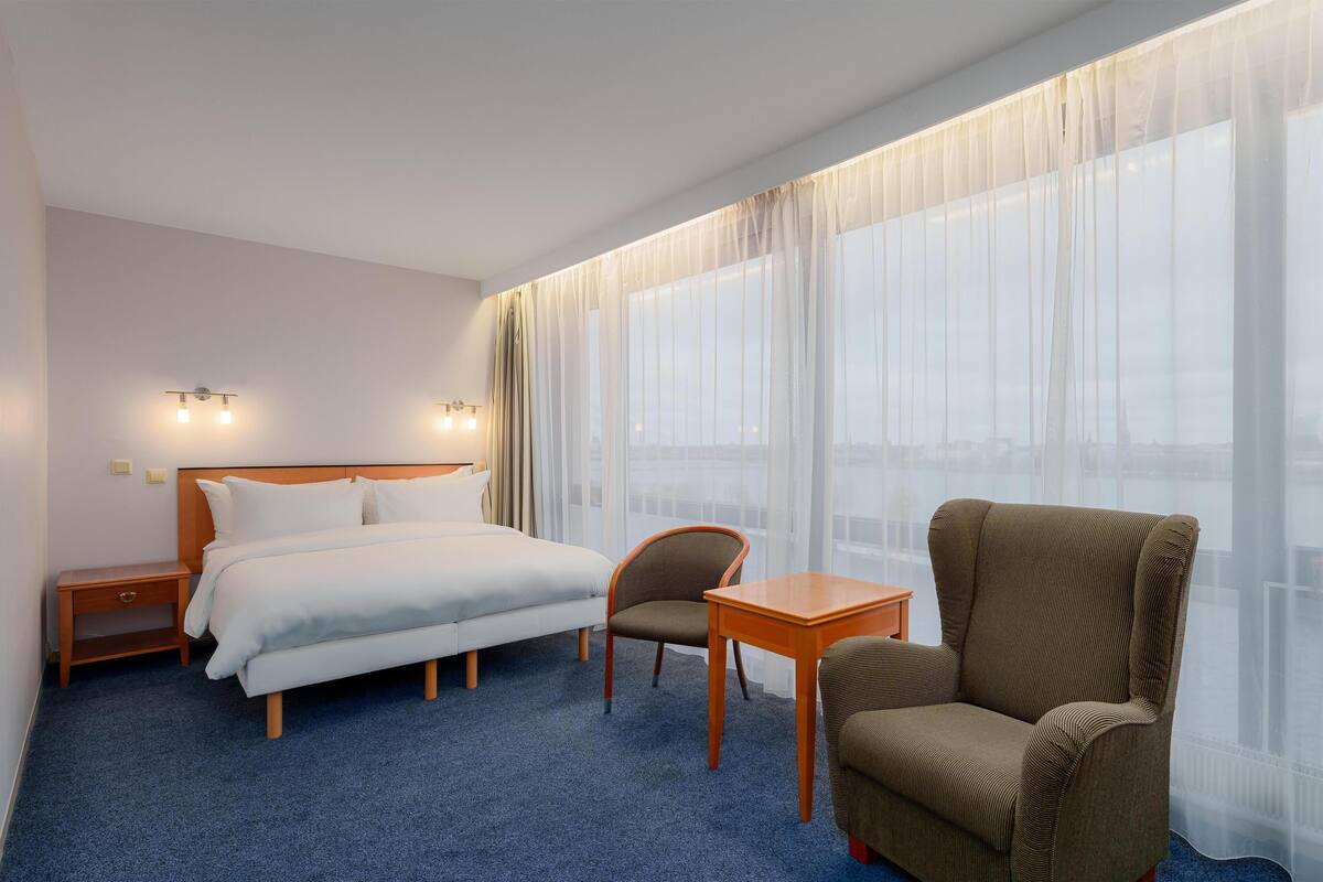 A hotel room with a bed, a chair, a small table, and a large window with sheer curtains.