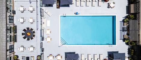 A rooftop pool area with a rectangular pool, multiple lounge chairs, and tables arranged around the pool.