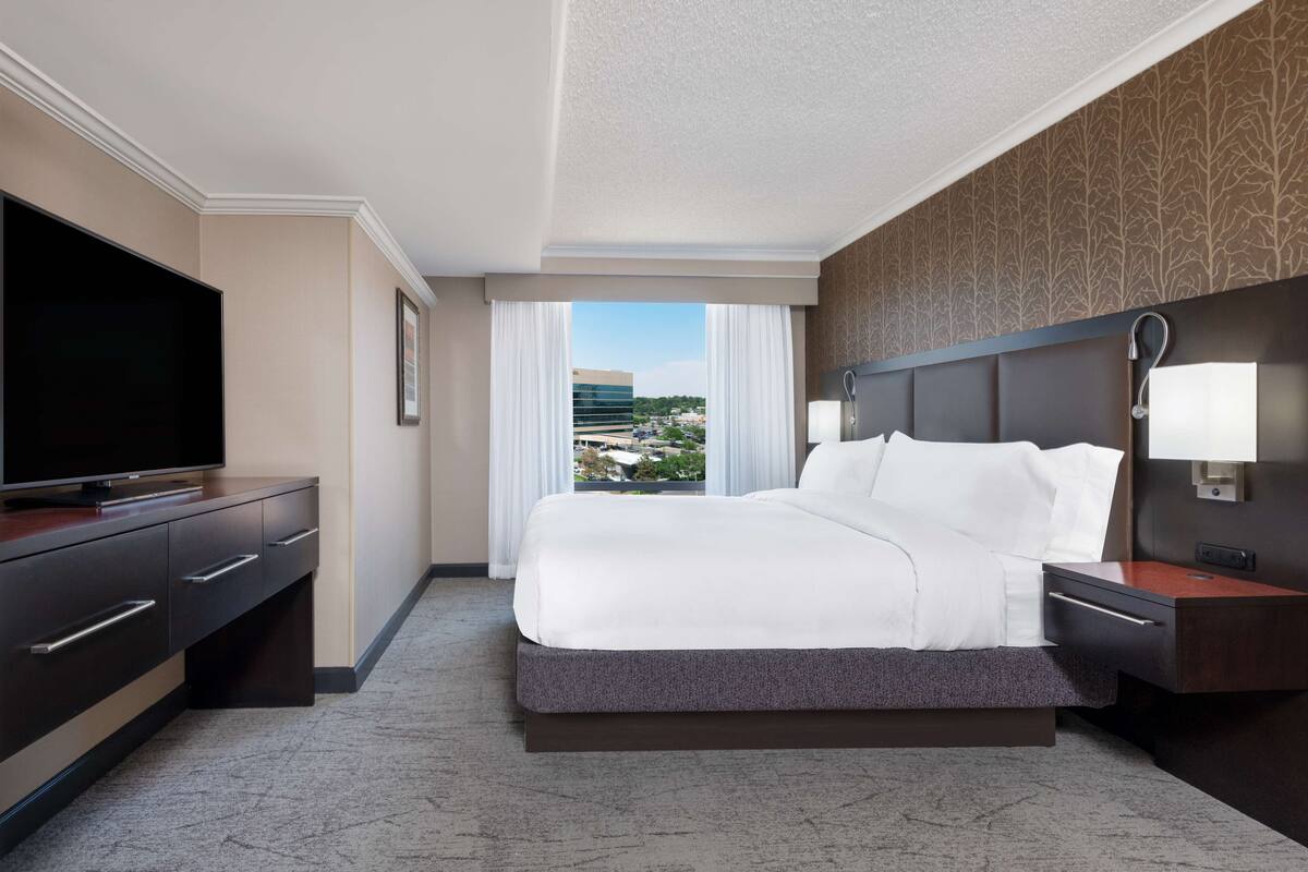 A hotel room with a large bed, a flat-screen TV, a desk, and a view of a cityscape through the window.