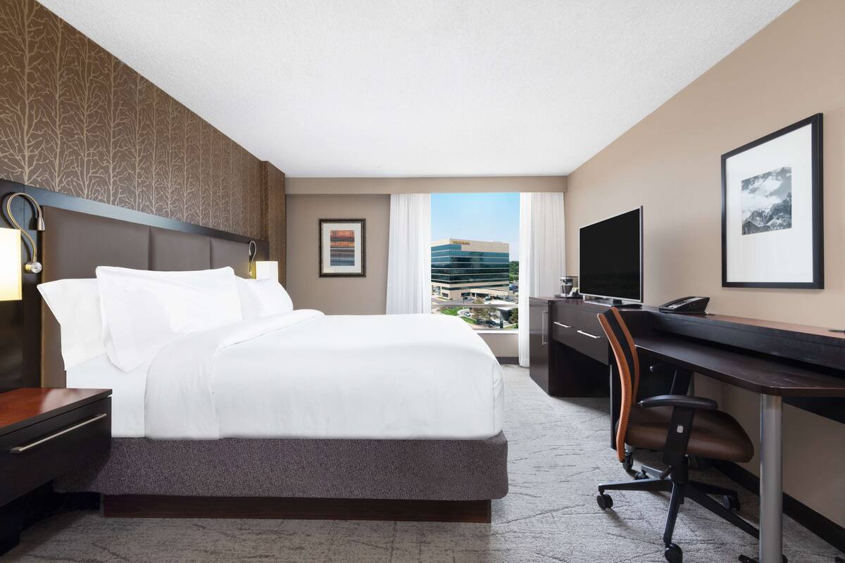 A hotel room with a bed, desk, chair, and a large window offering a city view.