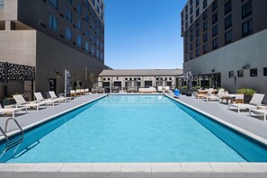 A rectangular pool with lounge chairs and umbrellas, surrounded by modern buildings with multiple windows. - DoubleTree by Hilton Denver Cherry Creek (Denver)