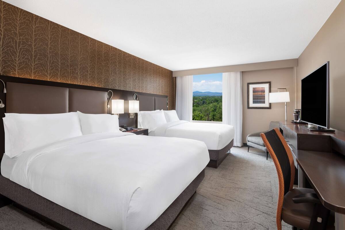 A hotel room with two beds, a desk, a chair, a TV, and a window with a view of greenery.