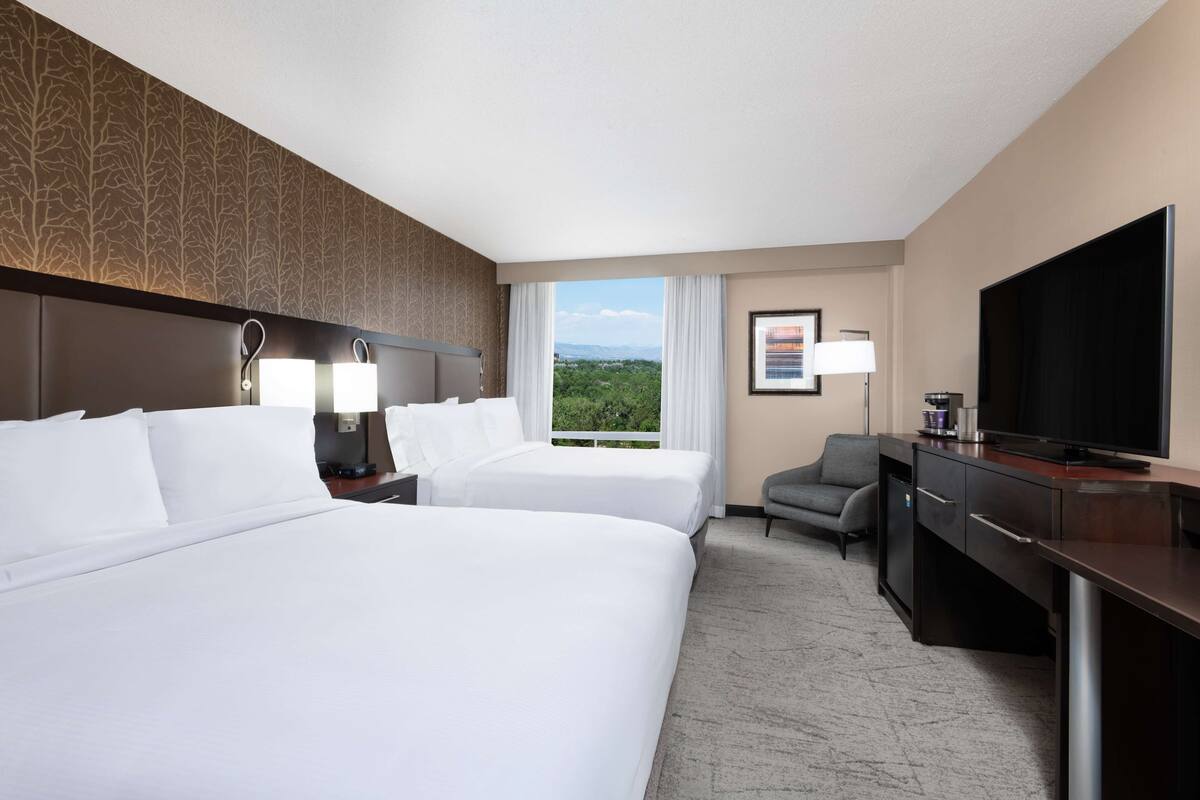 A hotel room with two beds, a desk, a television, and a view of the outdoors.