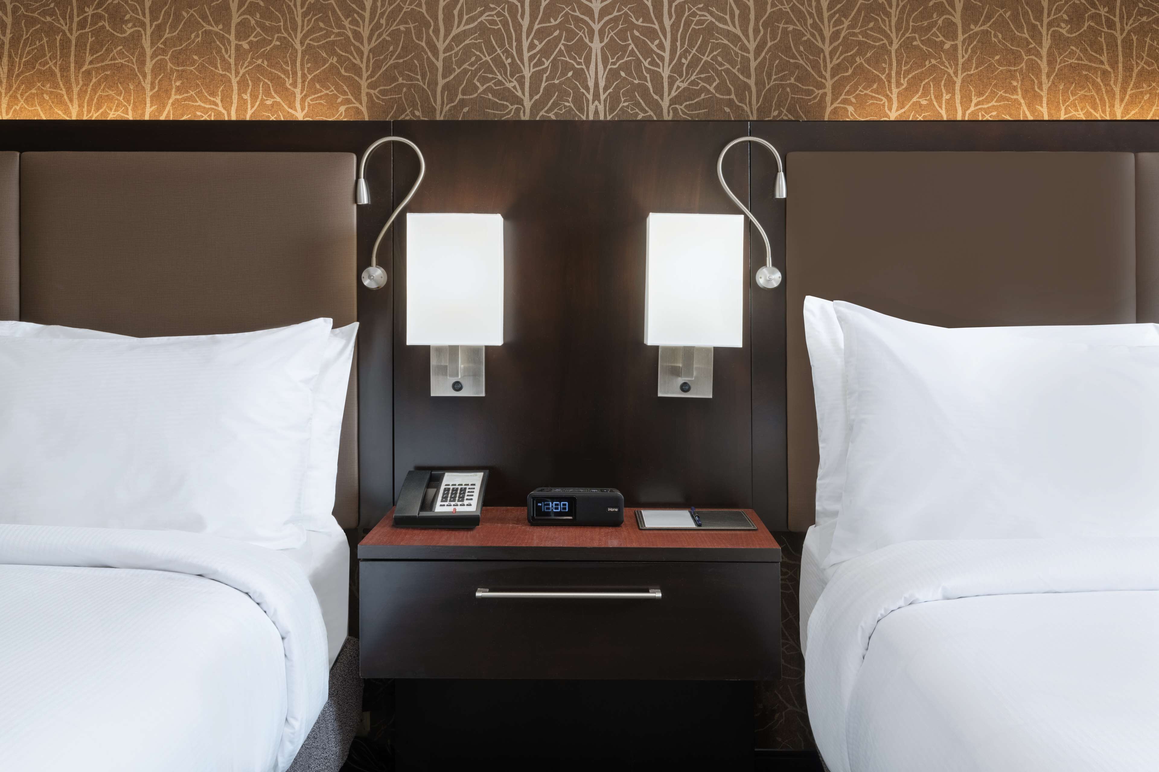 A hotel room with two beds, a bedside table with a phone, a digital clock, and wall-mounted lamps.