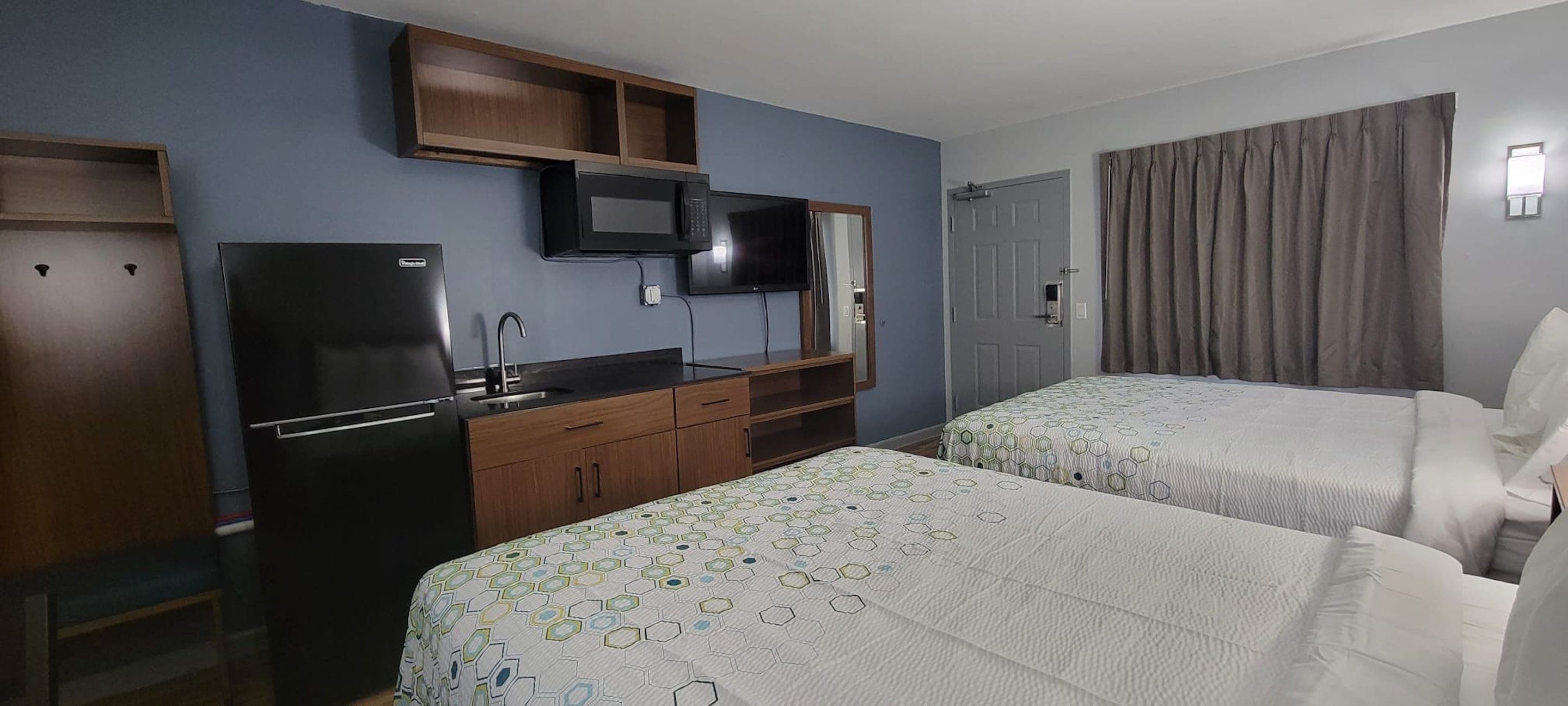 A hotel room with two beds, a kitchenette, a flat-screen TV, and a door with a curtain.