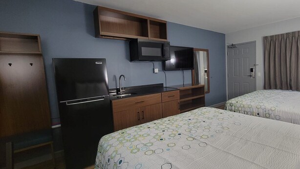A hotel room with two beds, a kitchenette, a flat-screen TV, and a door with a curtain.