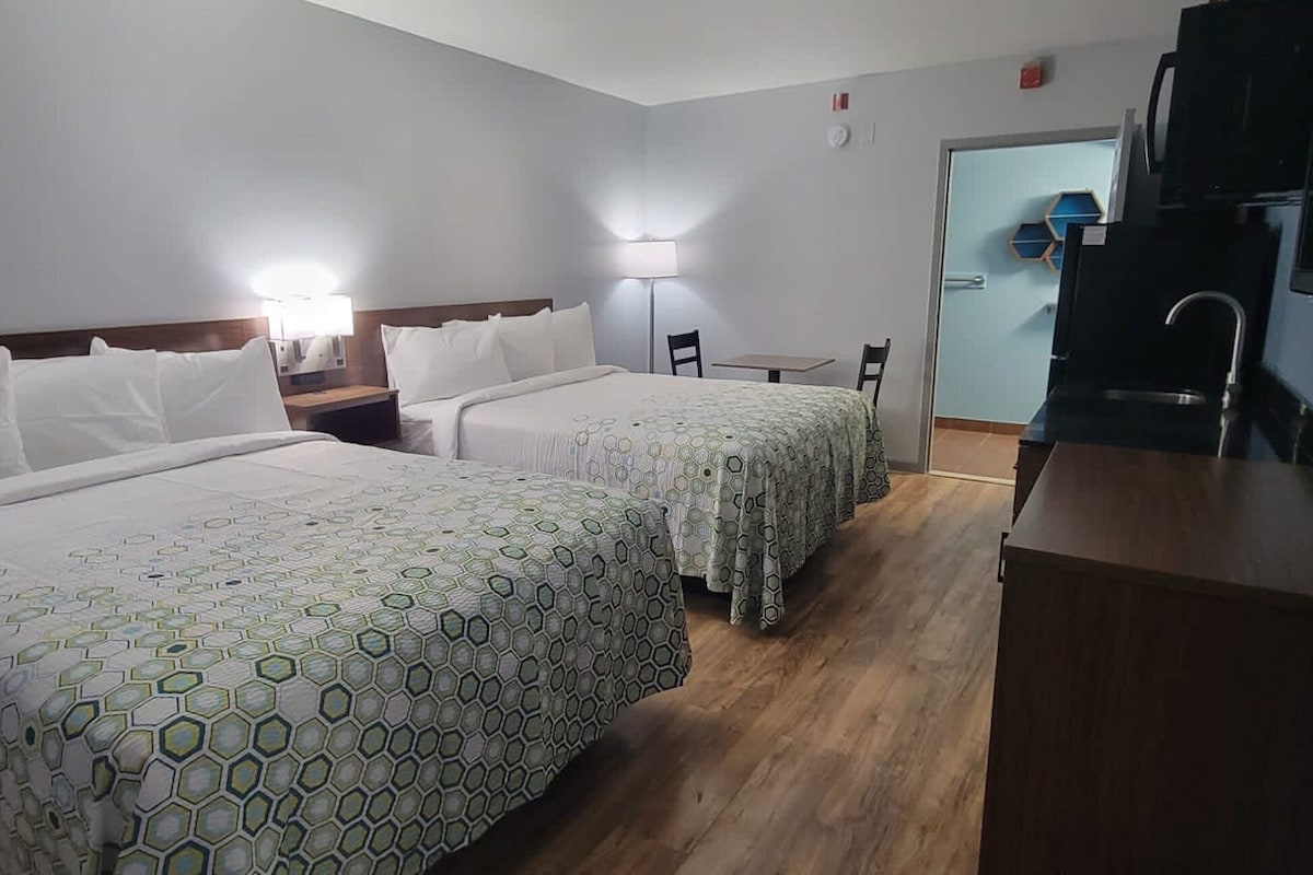 A hotel room with two beds, a small table, a chair, a TV, and a kitchenette.