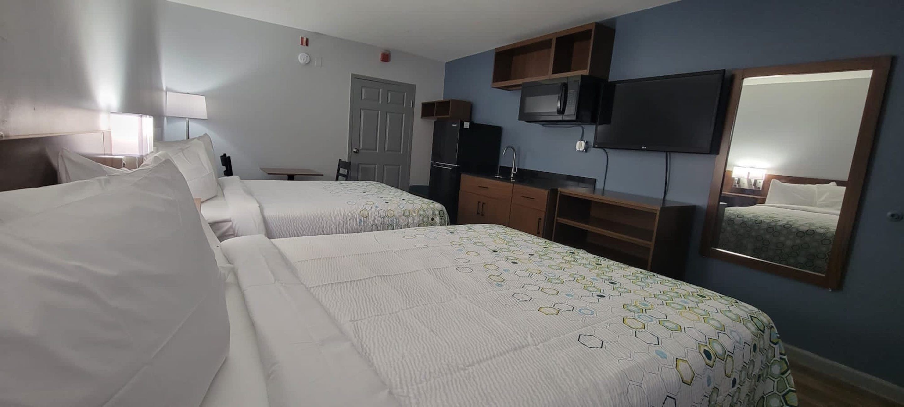 Habitación de hotel con dos camas, televisión, espejo y kitchenette.