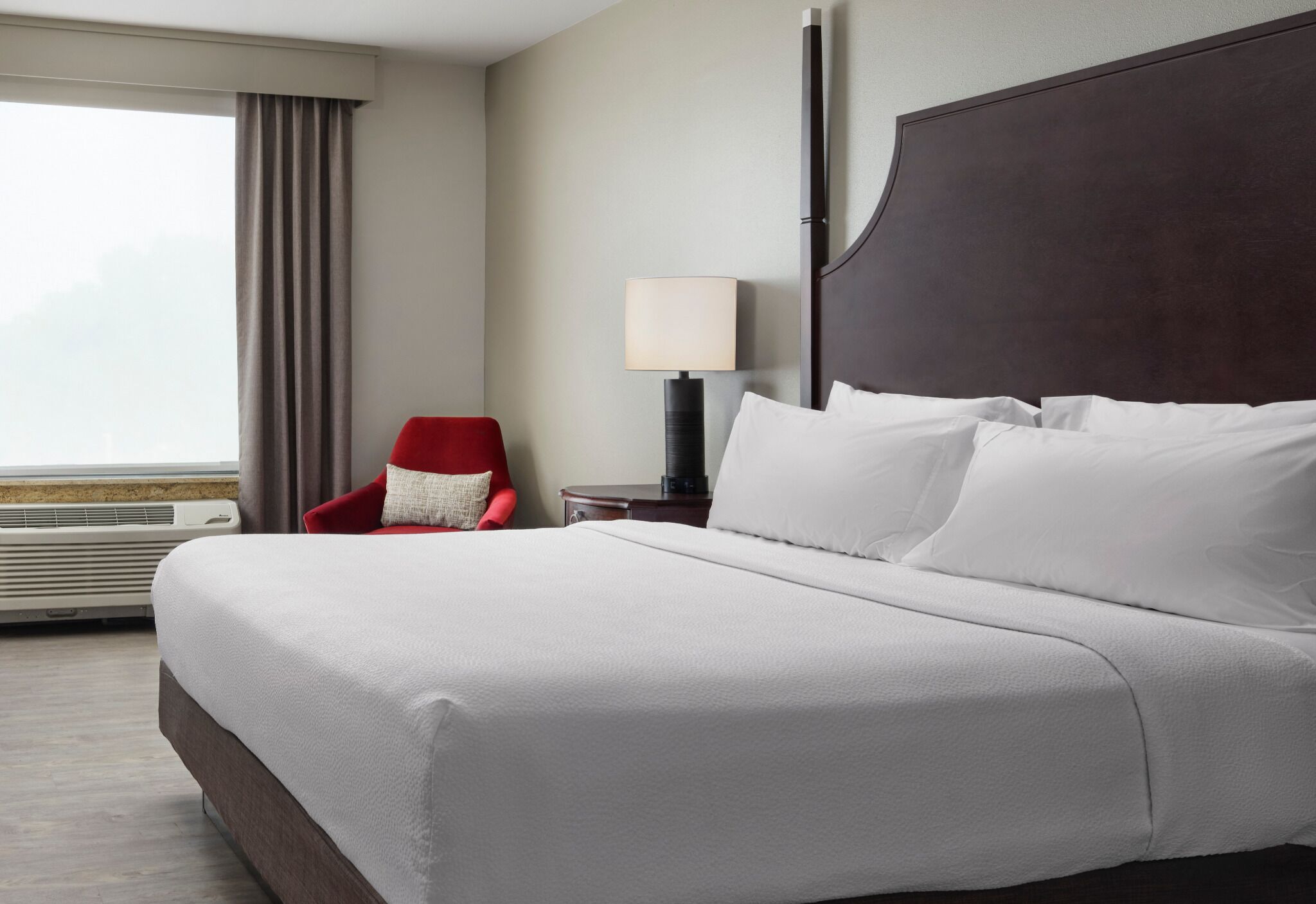 A hotel room with a large bed, a red chair, a bedside table with a lamp, and a view of the outdoors.