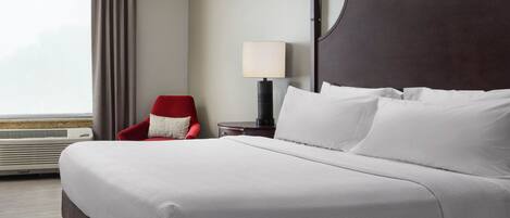 A hotel room with a large bed, a red chair, a bedside table with a lamp, and a view of the outdoors.