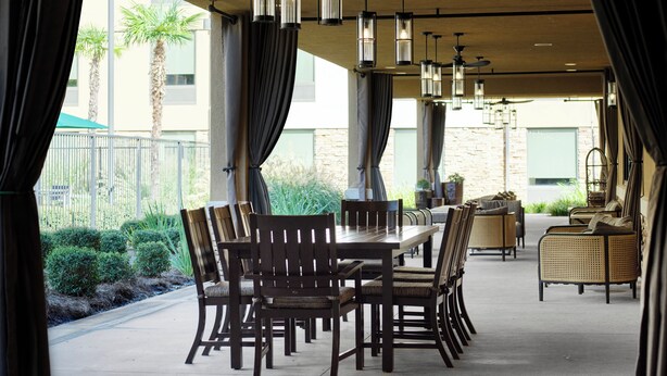 An outdoor dining area with a long table and chairs, surrounded by curtains and hanging lights.