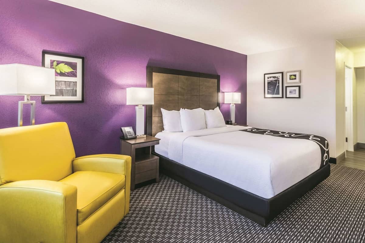 A hotel room with a bed, a yellow armchair, two bedside tables with lamps, and a wall with framed pictures.