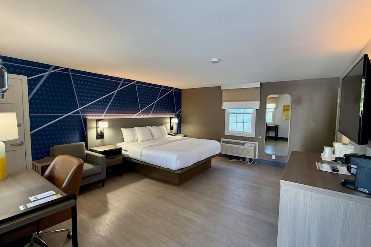 A hotel room with a bed, desk, chair, bedside tables, a TV, and a wall with geometric patterns.