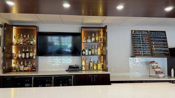 Un espace bar avec des étagères remplies de bouteilles d’alcool, un téléviseur et un tableau affichant la carte des boissons.