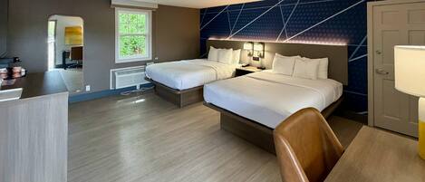 A hotel room with two beds, a desk, a chair, a desk lamp, a nightstand, a window, and a door.