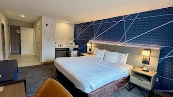 A hotel room with a large bed, a desk, a chair, a microwave, a mini-fridge, and a bathroom with a toilet and sink.