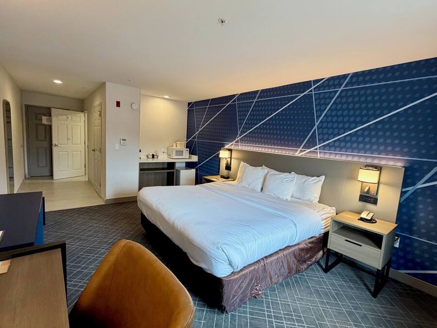 A hotel room with a large bed, a desk, a chair, a microwave, a mini-fridge, and a bathroom with a toilet and sink.