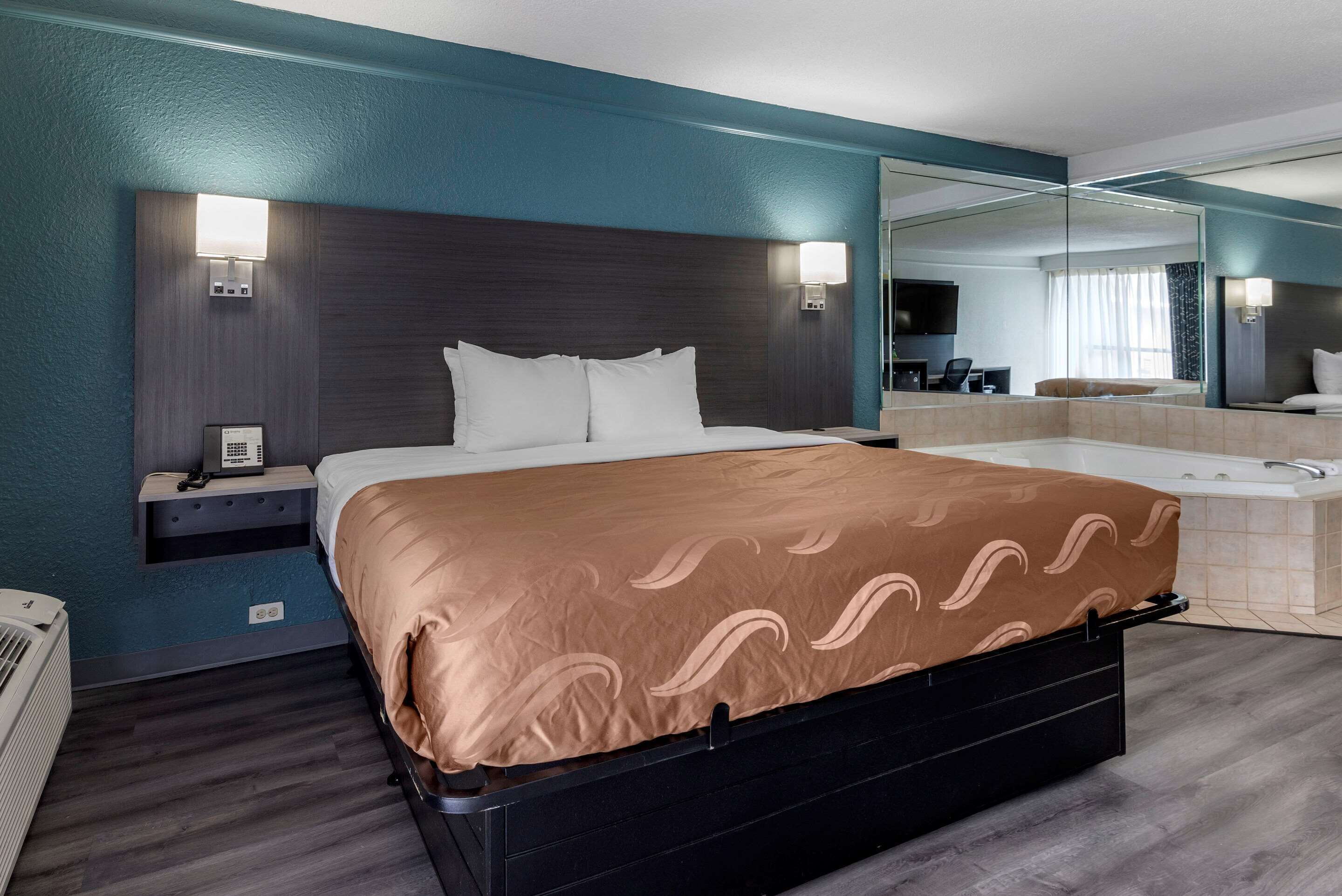 A modern hotel room with a large bed, a built-in TV, a bedside table, and a bathroom with a bathtub.