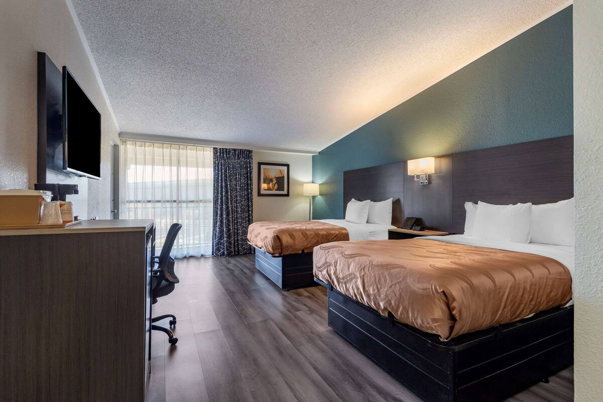 A hotel room with two beds, a desk, a TV, and a window with curtains.