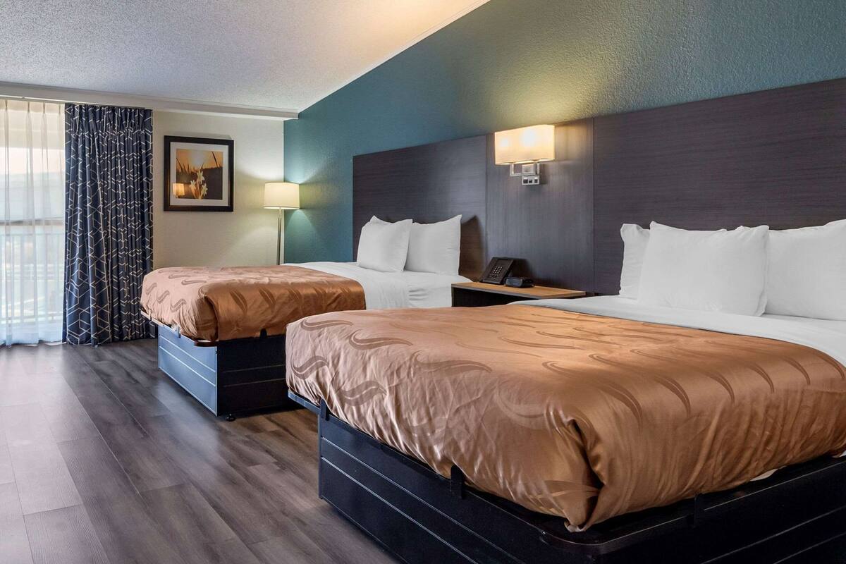 A hotel room with two beds, a nightstand with a phone, a wall-mounted lamp, and a window with curtains.