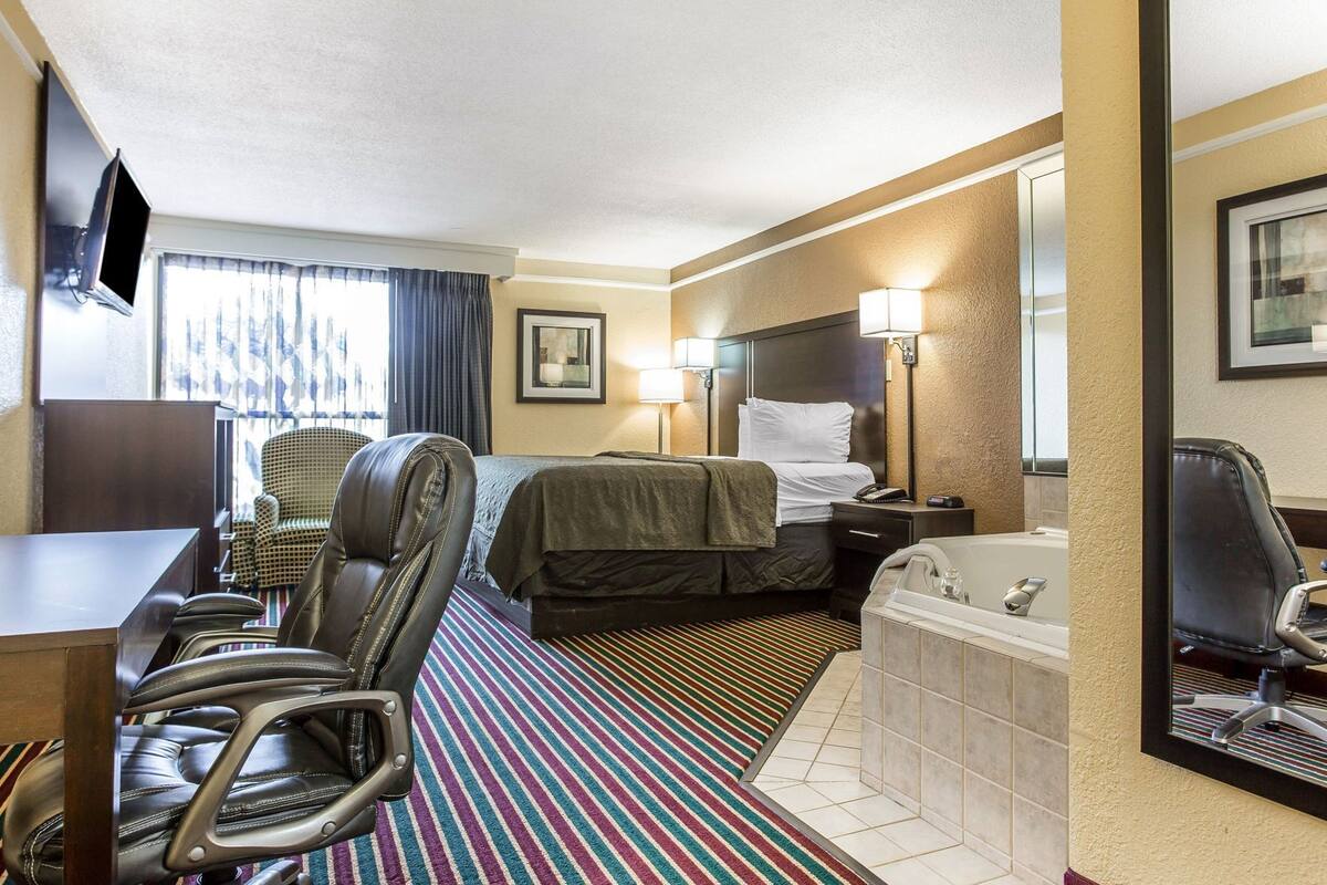 A hotel room with a bed, a desk, a chair, a desk lamp, a bedside table with a lamp, a window with curtains, a striped rug, and a bathtub.