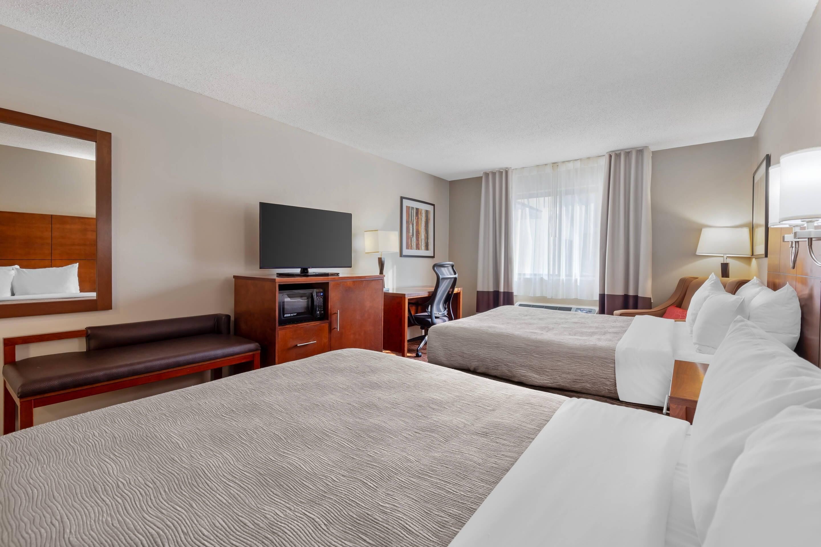 Habitación de hotel con dos camas, un televisor, un escritorio con computadora, una silla y un ventanal con cortinas.