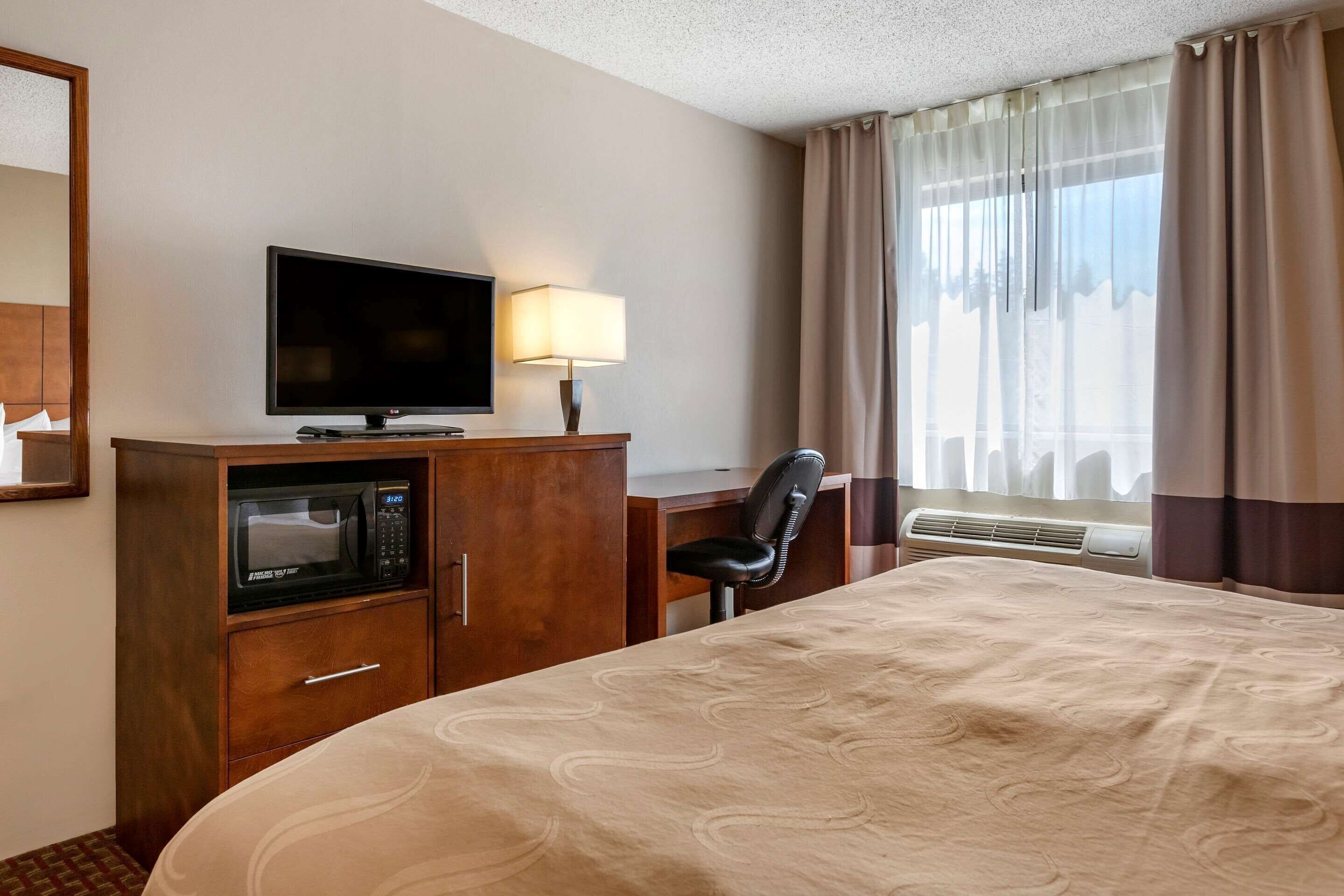 A hotel room with a bed, a TV, a microwave, a desk with a chair, and a window with curtains.