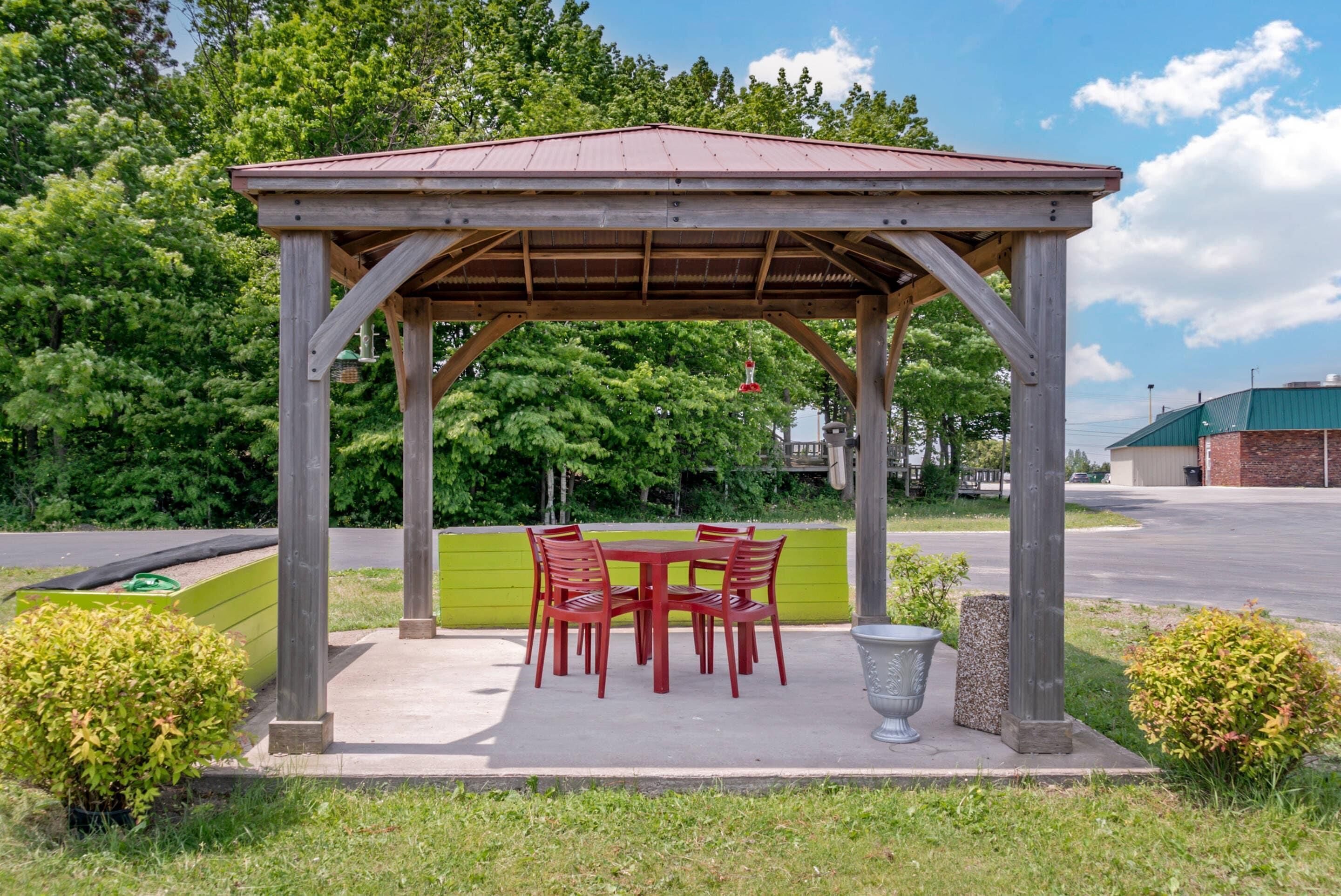 A wooden gazebo with a table and chairs, surrounded by greenery and a paved area.