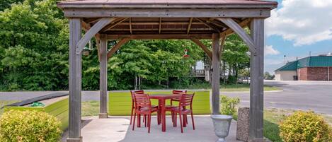 A wooden gazebo with a table and chairs, surrounded by greenery and a paved area.