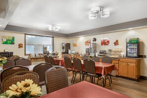 Un comedor con mesas y sillas, una zona de cocina con electrodomésticos, y un refrigerador con dispensador de bebidas.