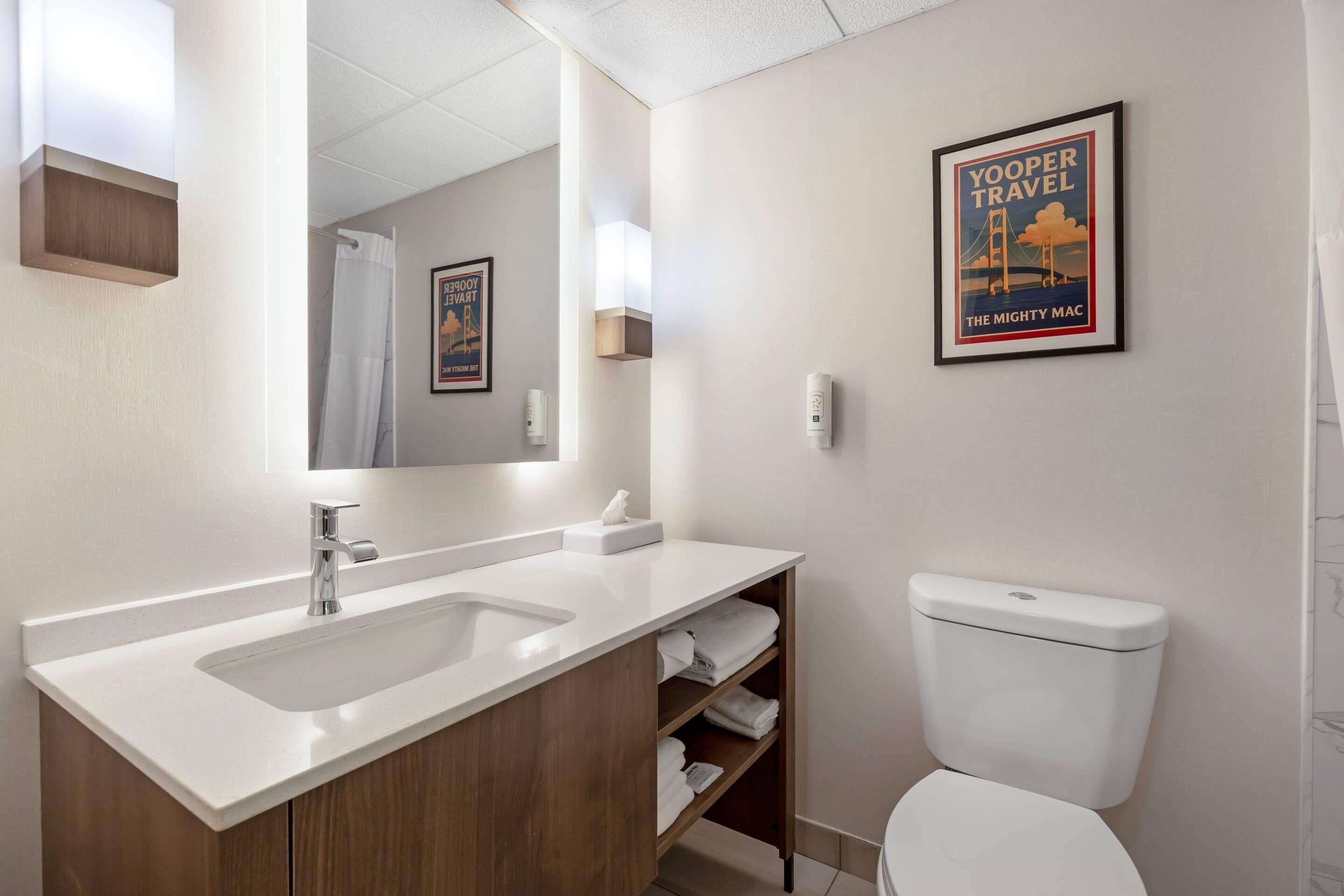 A modern bathroom with a white sink, mirror, and toilet. There are towels and a framed picture on the wall.