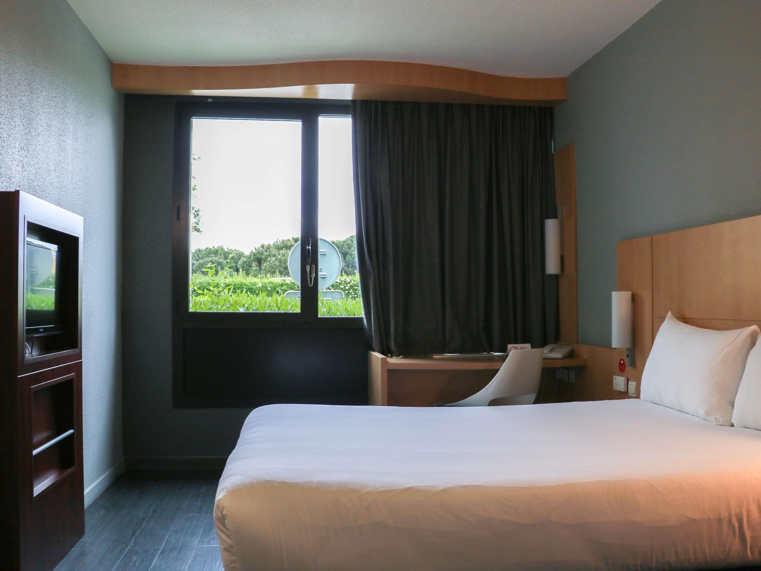 Habitación de hotel con cama, escritorio, silla, televisor y ventana con cortinas.