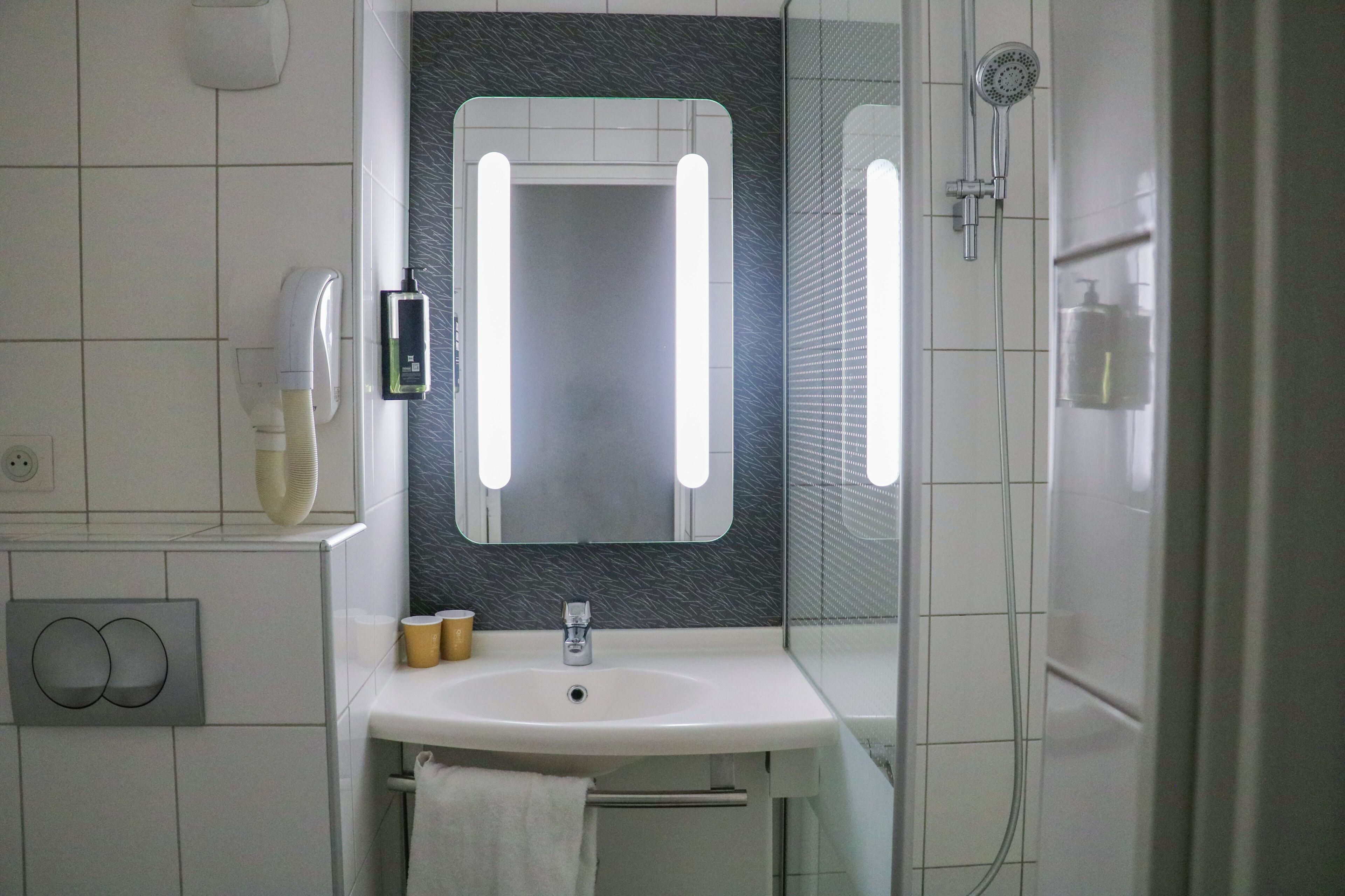 Un baño con lavamanos blanco, espejo con iluminación, cabezal de ducha de mano y dispensador de jabón.