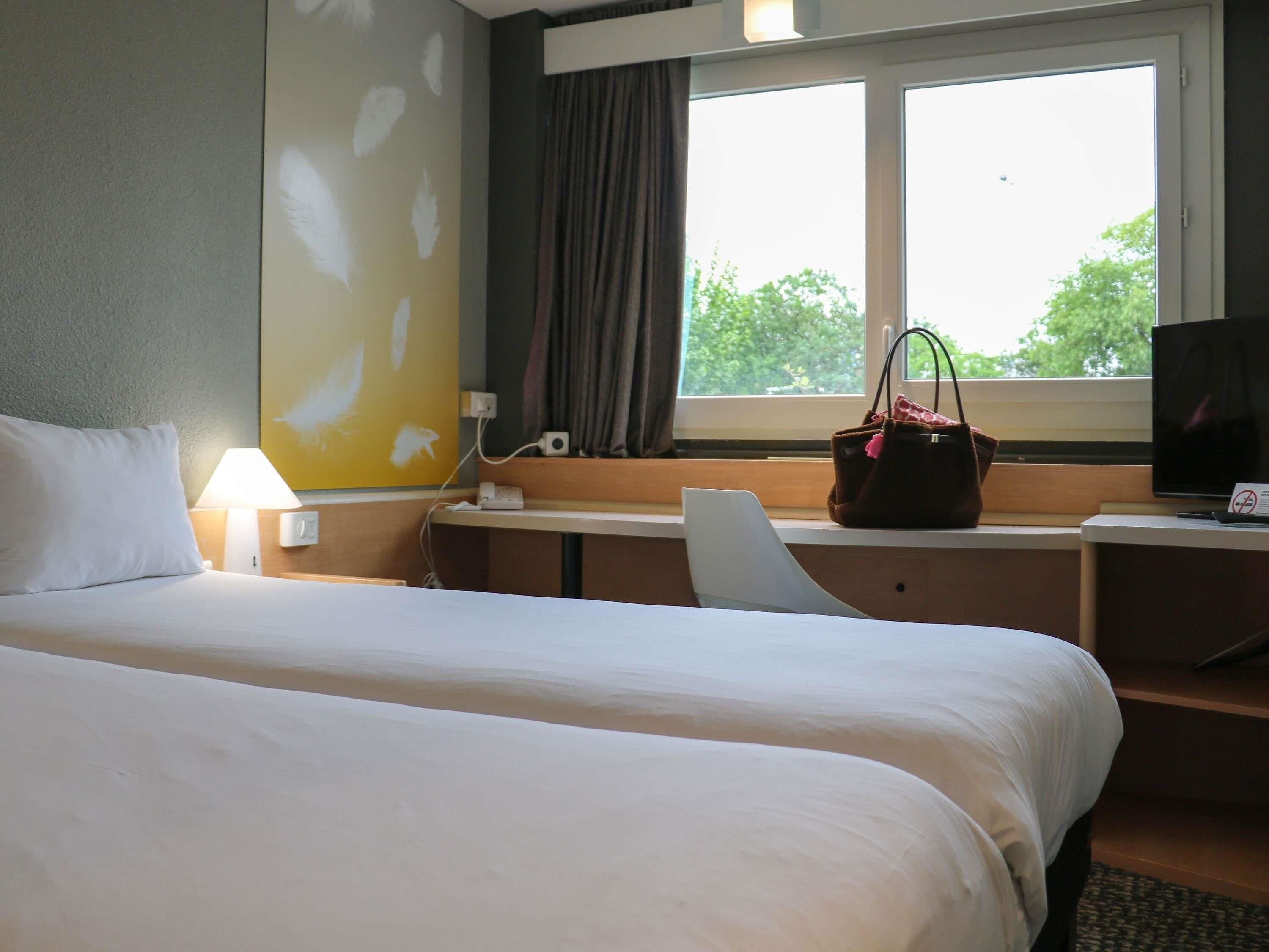 A hotel room with two beds, a desk with a computer, a TV, and a window with a view of trees.