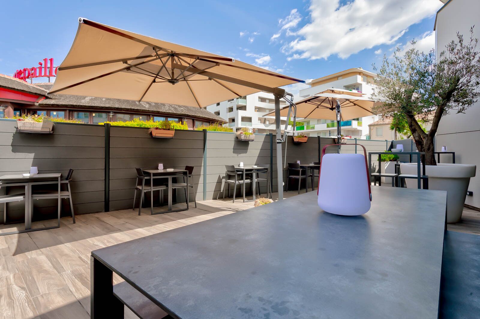 Un patio al aire libre con mesas y sillas, un paraguas grande y un edificio con un letrero al fondo.