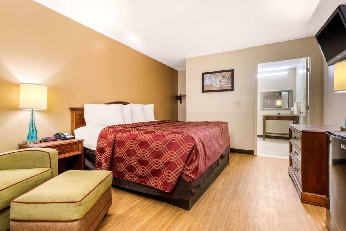 A hotel room with a bed, a desk, a chair, a TV, and a bathroom visible through an open door.