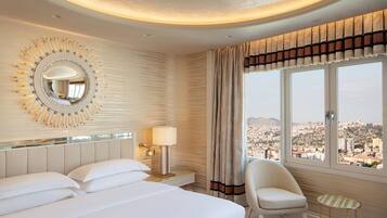 Ein Hotelzimmer mit einem großen Bett, einem Sessel, einem kleinen Tisch, einer Lampe, einem Spiegel und Blick auf die Stadt.