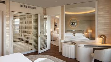 A modern hotel room with a large mirror, a curved desk, a comfortable chair, a bed, and a walk-in closet.