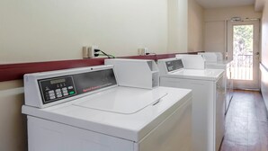 A row of commercial washers with control panels, a red baseboard, and a door leading to an outdoor area. - Red Roof Inn Atlanta - Suwanee/ Mall of Georgia (Suwanee)