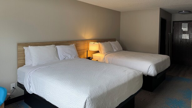 Quarto de hotel com duas camas, uma luminária de cabeceira, um painel de madeira, um tapete azul estampado e uma porta.