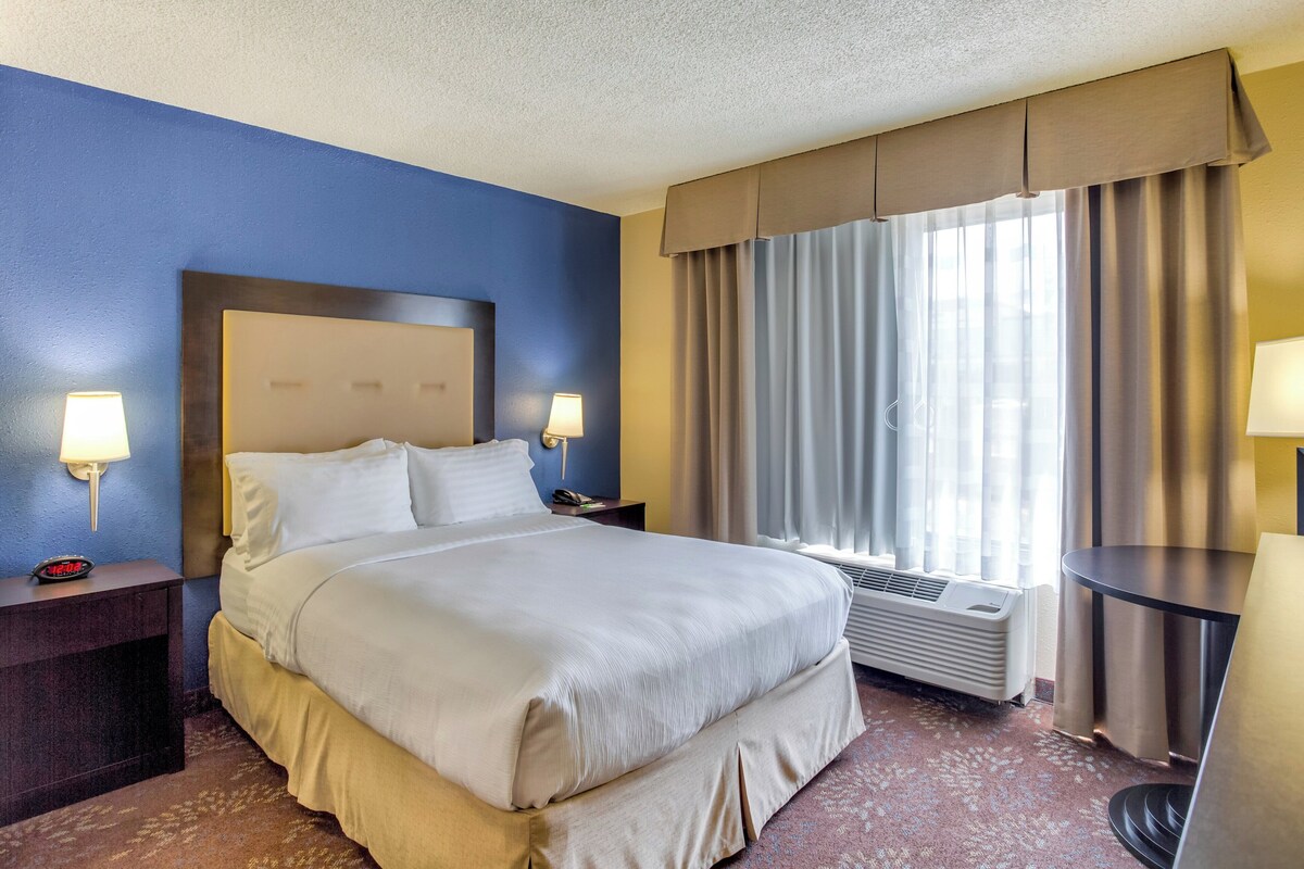 A hotel room with a large bed, bedside lamps, a desk, and a window with curtains.